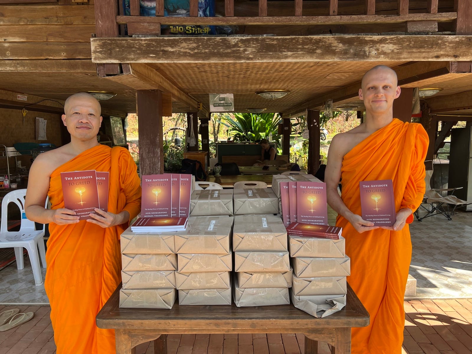 Ven. Nick and Ven. Michael holding copies of their books beside a table stacked with wrapped bundles of books.