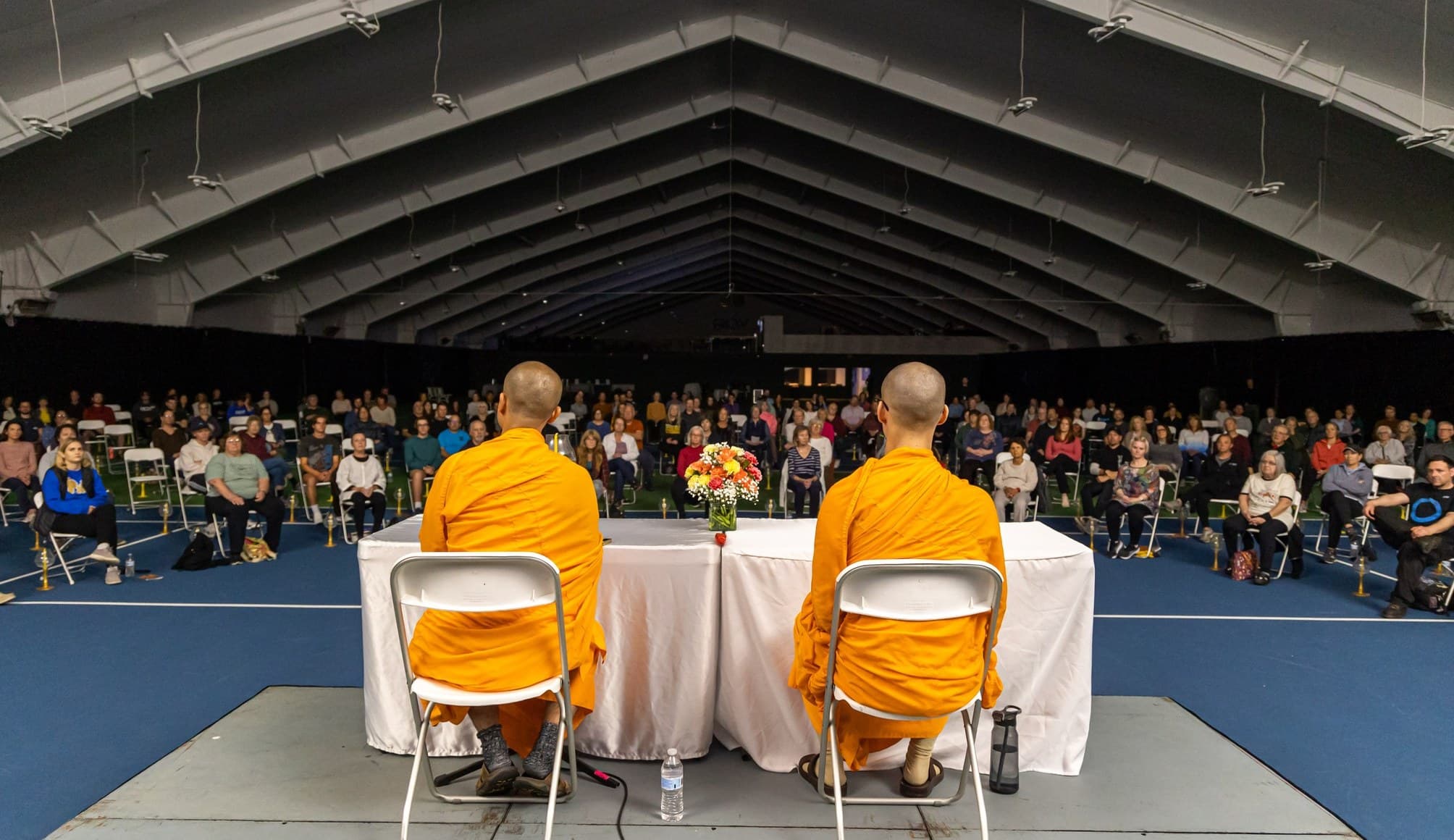 Two monks on stage before a large seated audience in a hall.