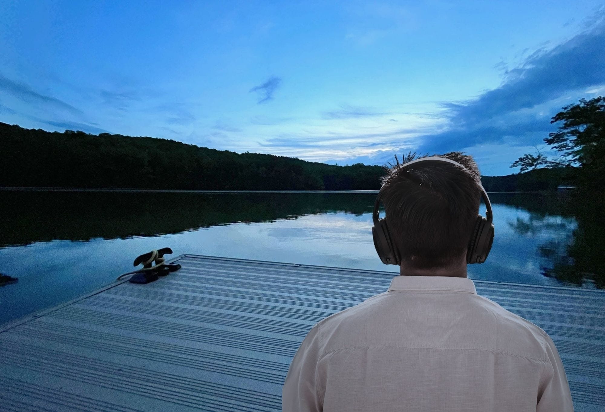Seen from behind, a person in headphones sits at the end of a dock at dusk, looking out over a still lake ringed by forest.