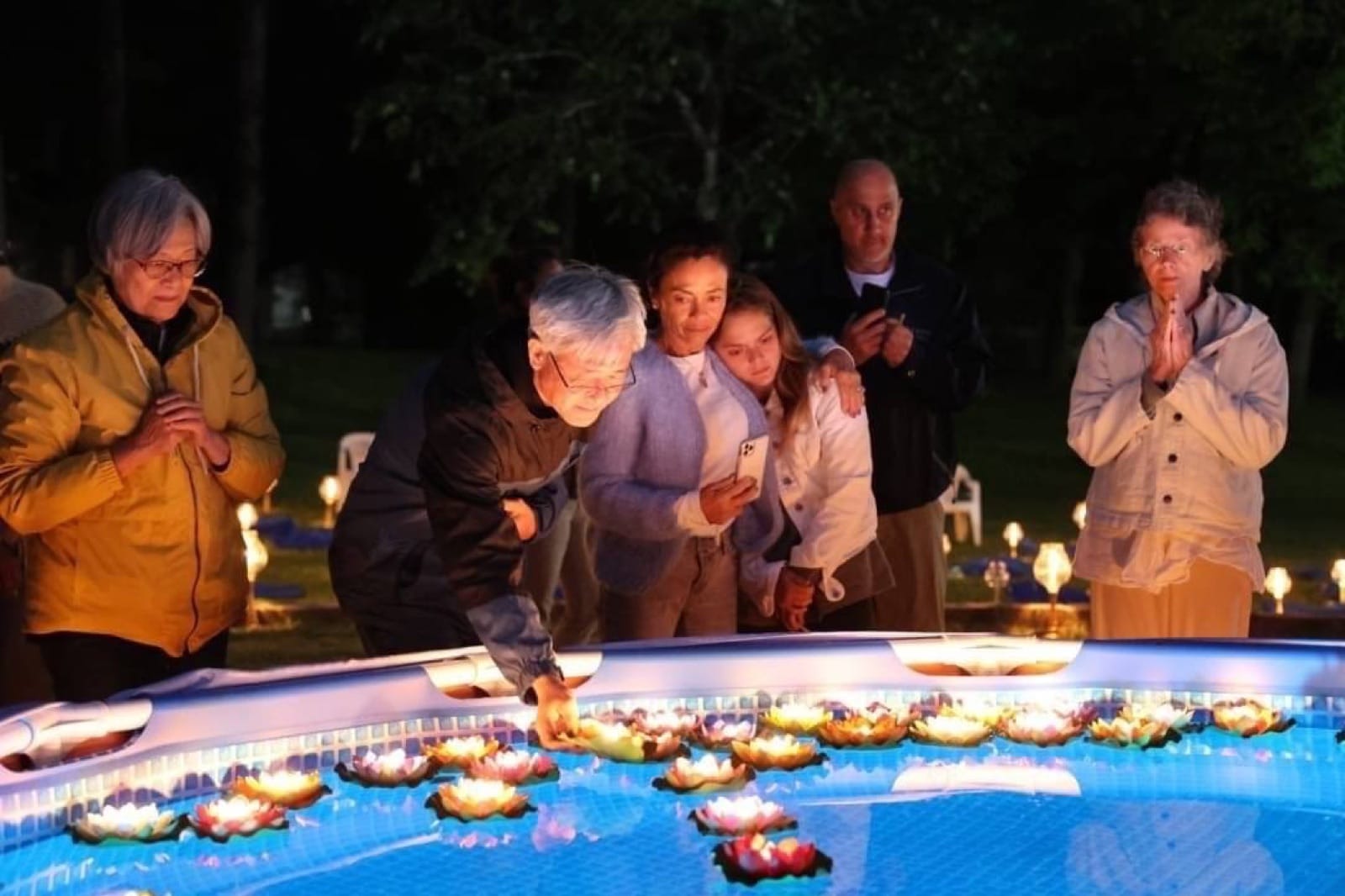People gathered at night around a pool of glowing lotus candle lanterns during a candlelight vigil.