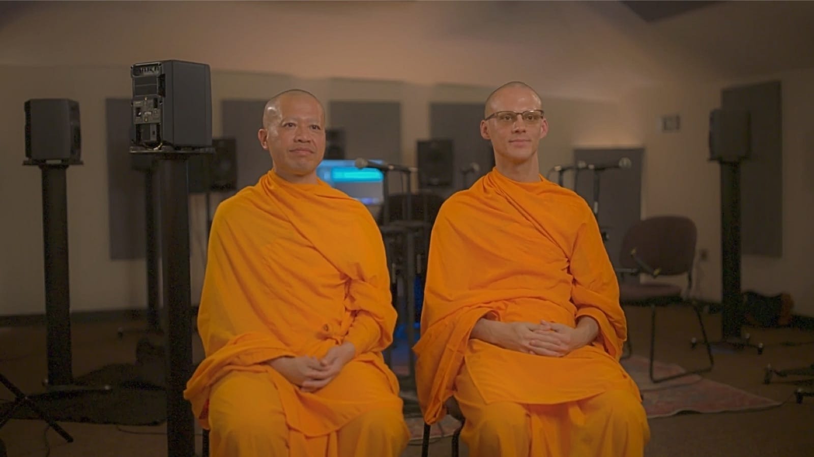 Two Buddhist monks in saffron robes seated calmly in a recording studio, surrounded by studio monitor speakers.